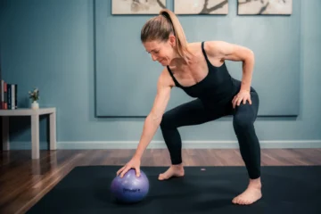 A woman performing a seated exercise on a yoga mat, using a therapy ball.