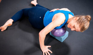 The woman uses a yoga block and ball under her chest with one hand on her back and one on the floor.
