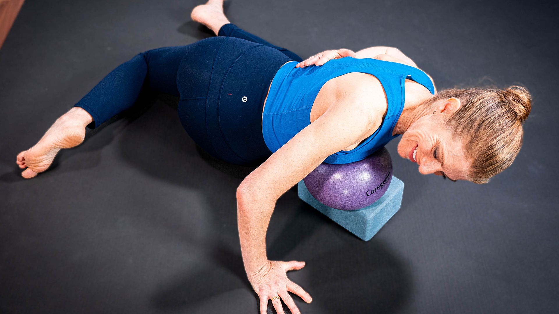 The woman uses a yoga block and ball under her chest with one hand on her back and one on the floor.