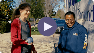 A woman and a man are smiling outdoors, with the woman holding a light purple ball.