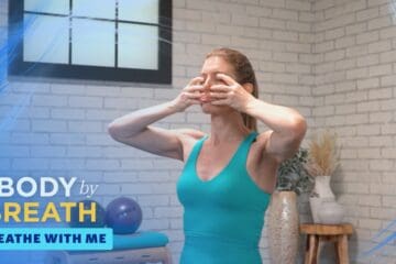 A woman stands in a turquoise sports bra, her hands positioned near her face, as if performing a facial exercise or yoga pose.