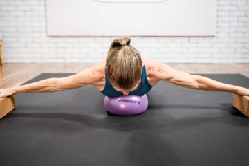 A woman is lying on a dark-gray yoga mat, her arms extended straight out to the sides, supporting the weight of her body.