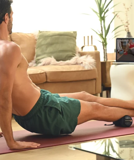 A man on a yoga mat in a living room, performing an exercise while following an online fitness class on a tablet