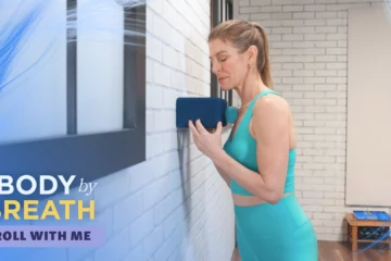 A woman poses in front of a wall featuring the words "body breath roll with me," symbolizing connection and physical awareness.