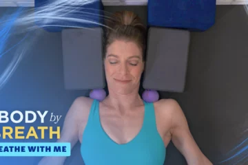 A woman lies on a dark gray mat with two gray yoga blocks under her head and shoulders, and purple balls placed on her shoulders.