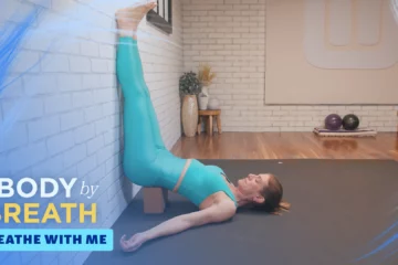A woman is lying on a dark gray yoga mat, her legs raised against a white brick wall.