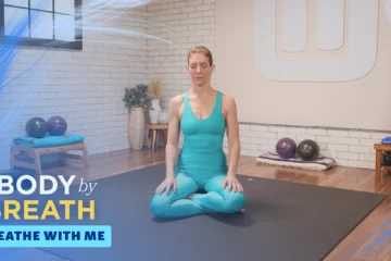 A woman sits in a seated meditation posture on a dark gray yoga mat in a light-filled room.