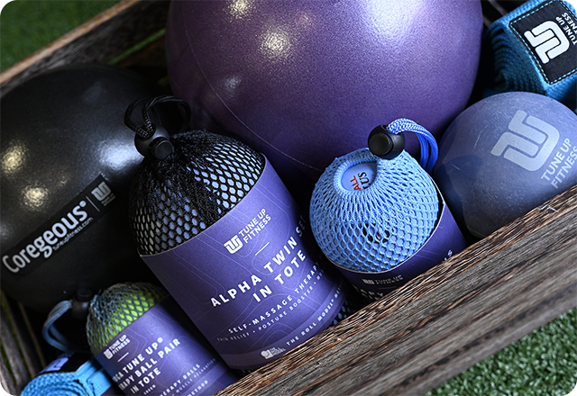 TUF_Bundled_Product_Lifestyle_Photo A wooden crate filled with Tune Up Fitness self-massage and recovery tools, including Alpha Twin massage balls in mesh pouches, a Coregeous fitness ball, and other purple and blue exercise balls, arranged on green turf.