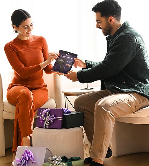S_Hero_Carousel_First Set_Left A smiling couple seated indoors exchanging a gift, with wrapped presents and bows arranged around them.