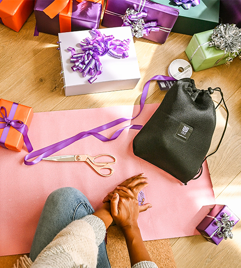 S_Hero_Carousel_First Set_Right Person wrapping gifts on a wooden floor, surrounded by colorful wrapped presents with bows, ribbon, scissors, and a black drawstring gift bag.