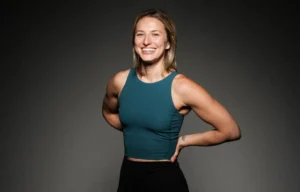 Smiling woman in a teal sleeveless top standing against a dark gray studio background.