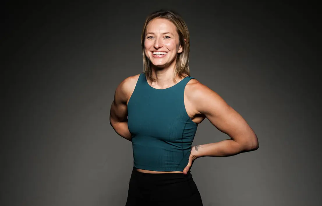 Smiling woman in a teal sleeveless top standing against a dark gray studio background.