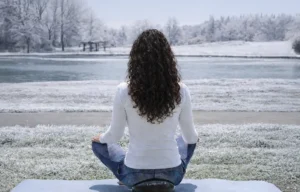 Woman seated cross-legged outdoors on a mat, facing a snowy lakeside landscape in a calm meditation pose.