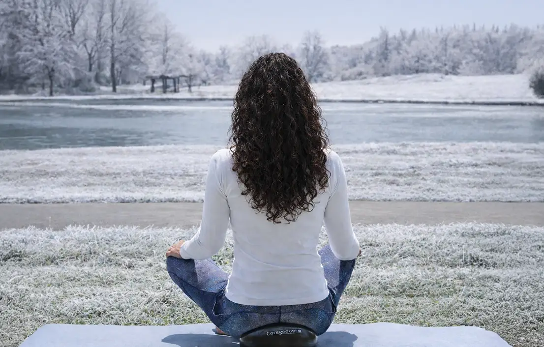 Woman seated cross-legged outdoors on a mat, facing a snowy lakeside landscape in a calm meditation pose.