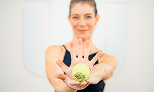 Homepage_MBR_Five_Fav_Hands-1 Woman holding a green therapy ball toward the camera while extending one hand forward.