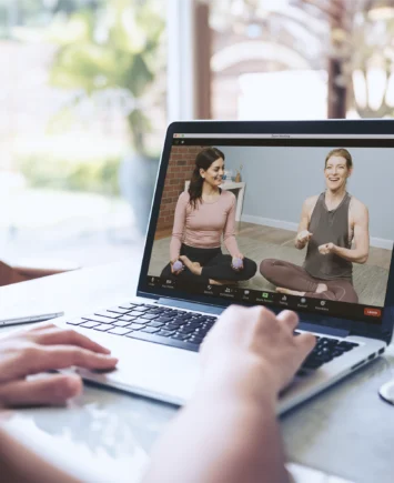 Proff-page-mbr1 Person watching an online movement class on a laptop, with two instructors demonstrating therapy ball exercises on screen.