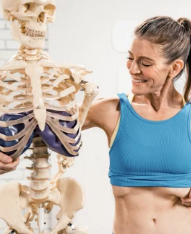 Proff-page-mbr2 Woman standing beside a skeleton model while demonstrating rib and torso anatomy with a purple therapy ball.