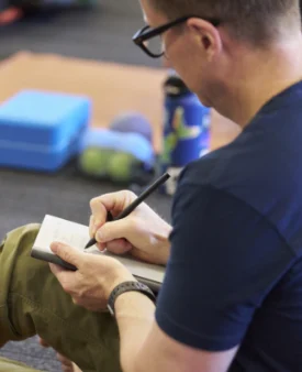 Proff-page-mbr4 Person seated on the floor writing notes in a small notebook, with fitness props and therapy balls nearby.