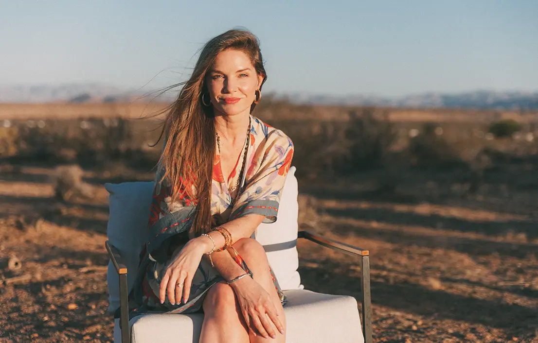 Ariel Kiley_Blog_Image_2 Woman seated outdoors on a white chair in warm sunlight, wearing a patterned robe with a blurred desert landscape in the background.